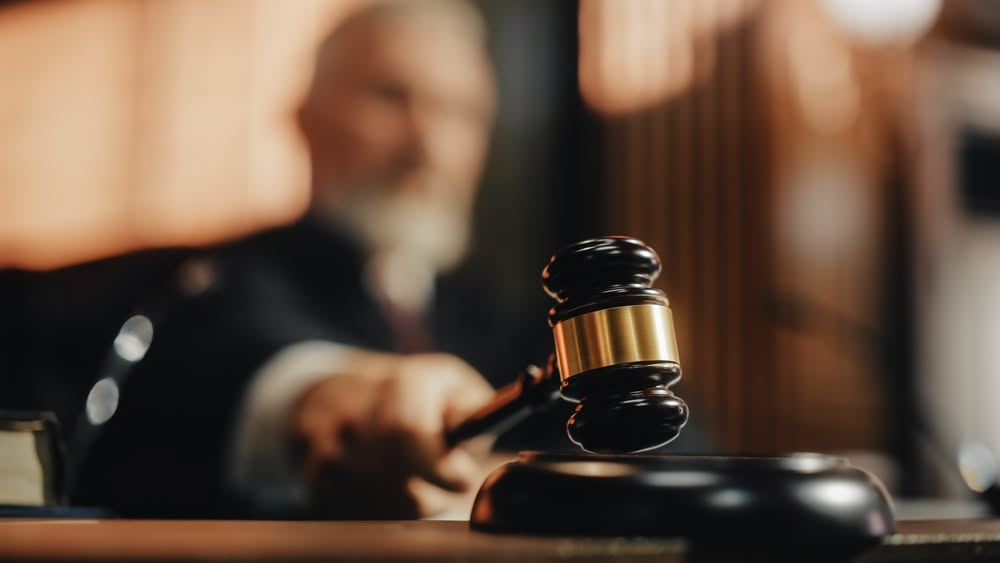 Martelo de juiz em primeiro plano sobre a mesa do tribunal, com o julgador desfocado ao fundo